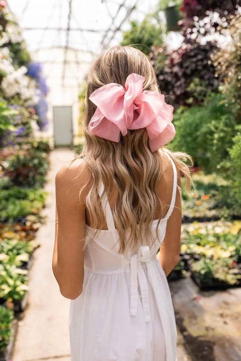 Woman in white dress with large pink hair bow, standing in a garden, Love Classic style
