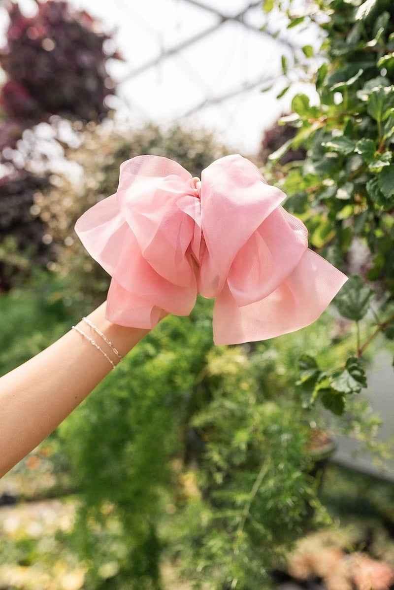 Hand holding a large pink hair bow from Love Classic, outdoors among green plants
