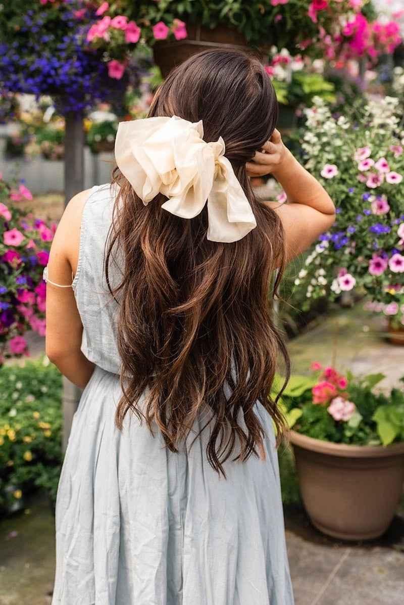 Woman with long wavy hair wearing a large cream bow in a garden with colorful flowers