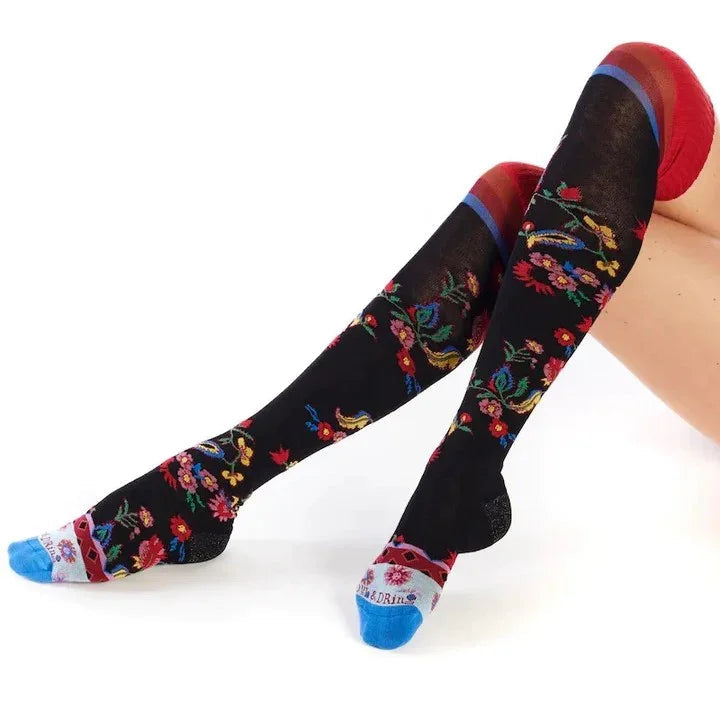 Black knee-high floral socks with colorful embroidery from Love Classic, worn by a seated model.