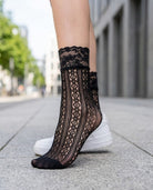 Floral Lace Vertical Fishnet Socks featuring a delicate openwork pattern with floral lace trim, worn with casual sneakers while standing on a city sidewalk, showcasing the stylish sheer texture, ankle length silhouette, and everyday fashion appeal.