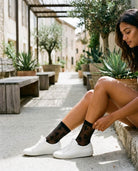 Floral Sheer Socks in Black featuring a lightweight see-through finish, soft floral pattern, and ankle cuff, styled with white sneakers while seated in a sunlit courtyard, highlighting casual everyday styling with subtle texture.