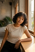 Striped Sleeveless Open Back Tank, cream sleeveless top with thin black horizontal stripes and a classic crew neckline, styled with dark high-waisted pants, shown on a model leaning by a sunlit window, highlighting the soft fabric and clean, minimal fit.