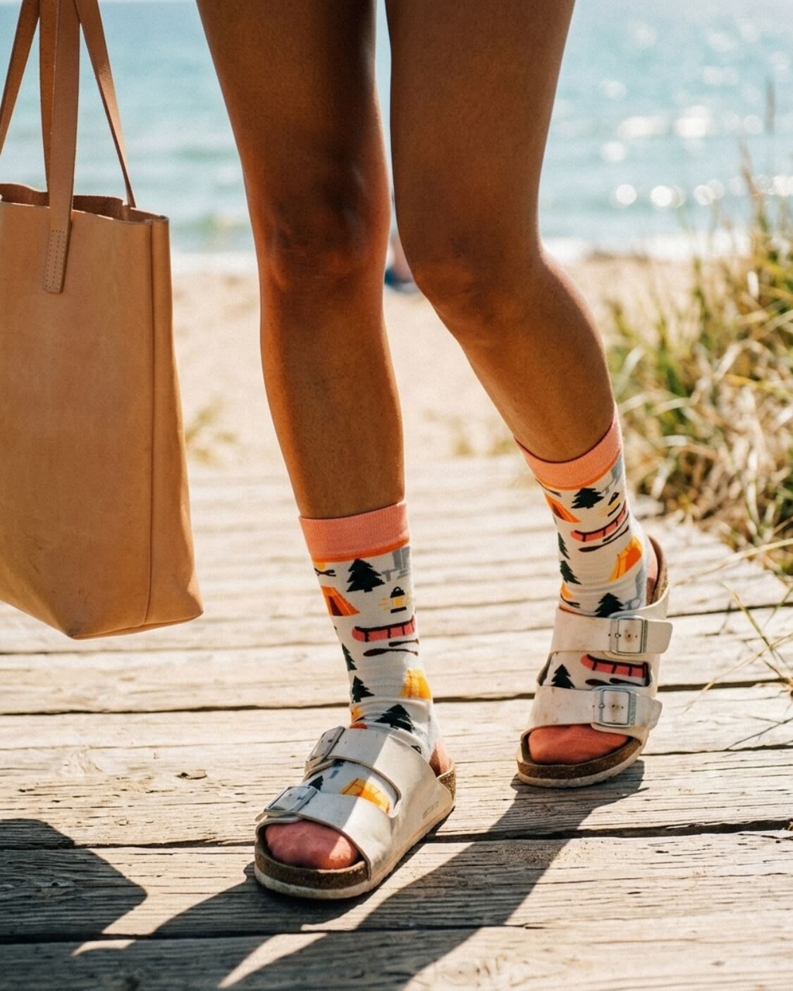 Crew Socks, Happy Camper featuring a colorful outdoor pattern with tents, trees, campfire graphics, and a soft ribbed cuff, worn with buckle sandals on a wooden beach walkway, highlighting playful casual styling for relaxed outdoor days.