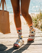 Crew Socks, Happy Camper featuring a colorful outdoor pattern with tents, trees, campfire graphics, and a soft ribbed cuff, worn with buckle sandals on a wooden beach walkway, highlighting playful casual styling for relaxed outdoor days.