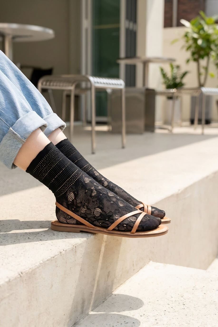Sheer Floral Lace Crew Socks in black with delicate floral lace pattern, styled with rolled cuff jeans and strappy flat sandals while seated on outdoor concrete steps, highlighting the semi-transparent texture and breathable knit.