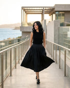 Poplin Cotton Smocked Flare Midi Skirt featuring a high waist, full flared shape, gathered front panel, and side pockets, worn with a sleeveless top on a coastal walkway, highlighting soft volume and easy movement.