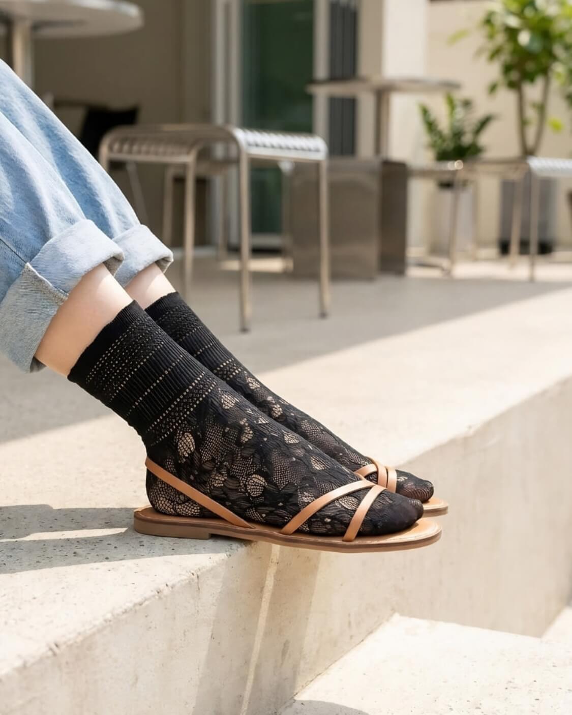 Sheer Floral Lace Crew Socks in black with delicate floral lace pattern, styled with rolled cuff jeans and strappy flat sandals while seated on outdoor concrete steps, highlighting the semi-transparent texture and breathable knit.