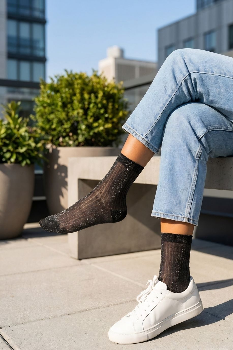 Sheer Metallic Ribbed Crew Socks in black with subtle shimmer, styled with light wash rolled cuff jeans and white sneakers while seated on a concrete bench, highlighting the semi-transparent ribbed texture and lightweight breathable fabric.