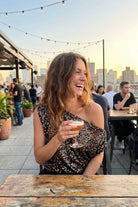 Premiere Night Top one shoulder sequin top styled at a rooftop bar setting during sunset, paired with wide leg pants, highlighting the relaxed fit and evening ready style.