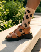 Sheer Argyle Crew Socks in beige and black with classic diamond pattern, styled with brown platform sandals while seated on outdoor steps, highlighting the semi-transparent fabric and modern argyle design.