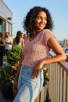Crochet Knit Cap Sleeve Sweater Top, blush pink open-knit sweater with cap sleeves and scalloped V-neckline, styled with light-wash high-rise jeans, worn by a model leaning on a balcony railing at a rooftop gathering, highlighting the airy texture and relaxed fit in warm sunlight.