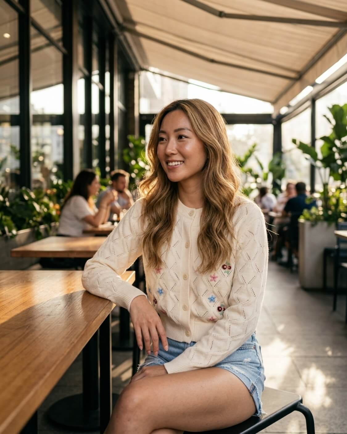 Pop Floral Sweater Weaved Cardigan featuring a button-front closure, open weave pattern, ribbed cuffs, and small floral embroidery, worn with denim shorts while seated in a bright café, highlighting soft texture and casual styling.