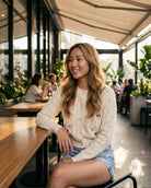 Pop Floral Sweater Weaved Cardigan featuring a button-front closure, open weave pattern, ribbed cuffs, and small floral embroidery, worn with denim shorts while seated in a bright café, highlighting soft texture and casual styling.