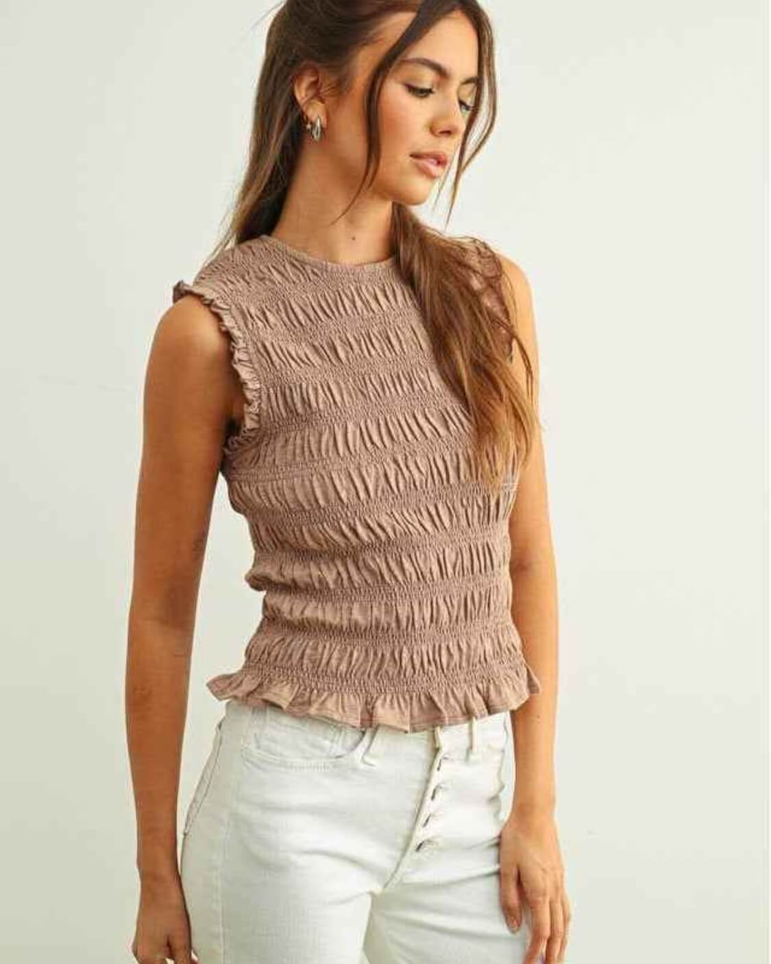 Sleeveless with All Over Smocking Top featuring a fitted all over smocked texture with soft ruffle sleeve trim and a ruffled hemline, styled with high waisted button front jeans in a side angled pose against a clean neutral backdrop, highlighting stretch comfort and flattering shape.