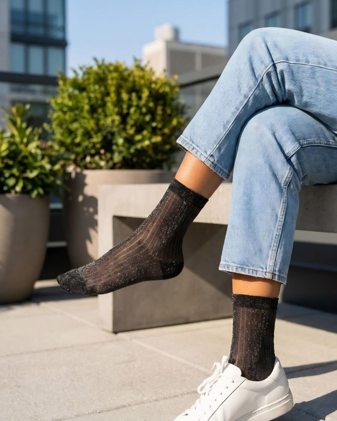 Sheer Metallic Ribbed Crew Socks in black with subtle shimmer, styled with light wash rolled cuff jeans and white sneakers while seated on a concrete bench, highlighting the semi-transparent ribbed texture and lightweight breathable fabric.