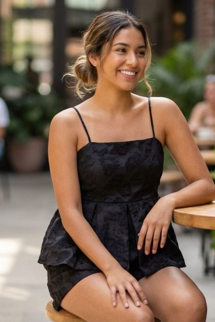 Sophie Top, black spaghetti strap peplum top with a tonal textured pattern, worn in a seated pose at a café table, emphasizing the structured bodice and soft volume at the waist.