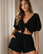 Lucia Shorts black two piece set featuring a tie front cropped top with puff sleeves and elastic waist shorts with pockets, styled indoors near a window with soft natural lighting, highlighting the flattering fit and relaxed everyday wear feel.
