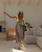 Metallic Fabric Pleat Detailed Skirt featuring a softly structured pleated front with luminous metallic texture and airy drape, styled in a sunlit living space with pointed heels, highlighting graceful flow and elevated everyday wear.