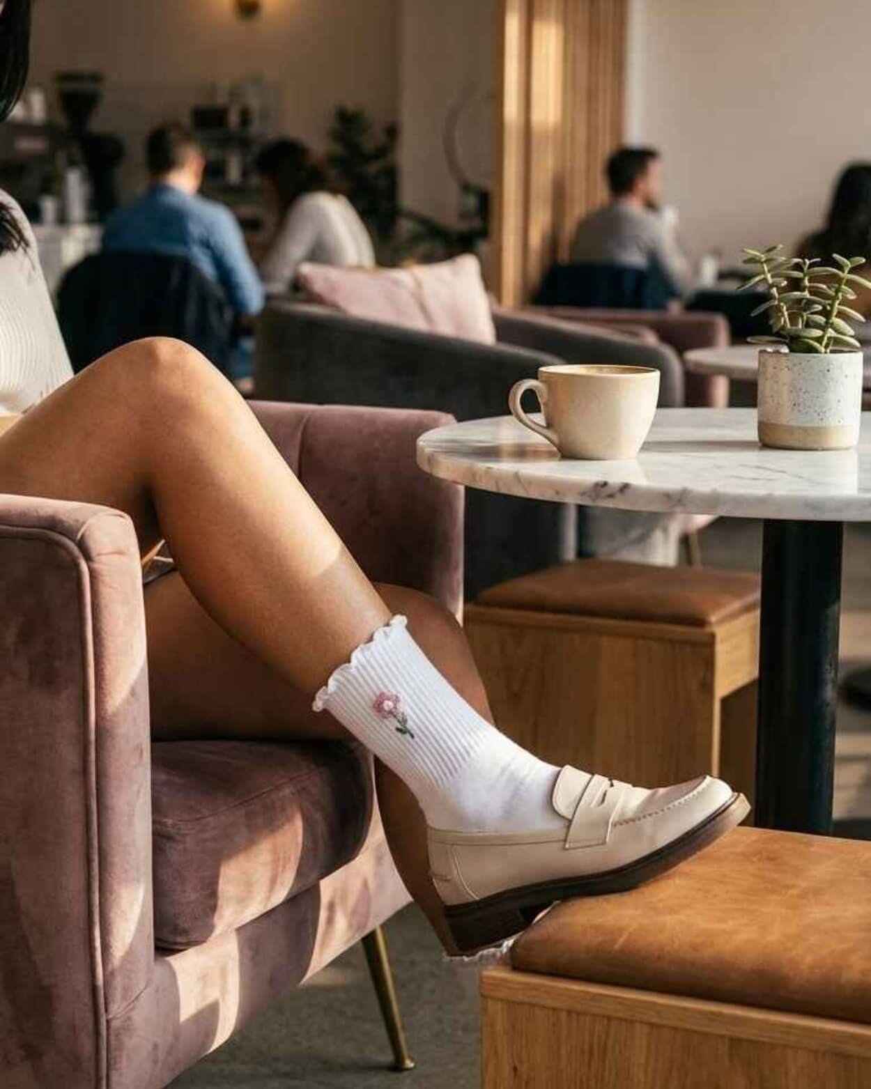 3-Pack Blush Pearl Rose Ruffle Crew Socks featuring a ribbed crew sock with a ruffled cuff and small flower embroidery, worn with a loafer while relaxing in a café lounge setting, highlighting casual styling, soft comfort, and a polished everyday finish.