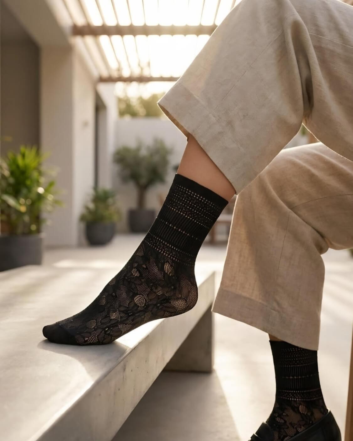Sheer Floral Lace Crew Socks in black with intricate floral lace design, worn with tailored trousers and black loafers while sitting on a sunlit bench, showcasing the lightweight, sheer finish and elegant pattern.