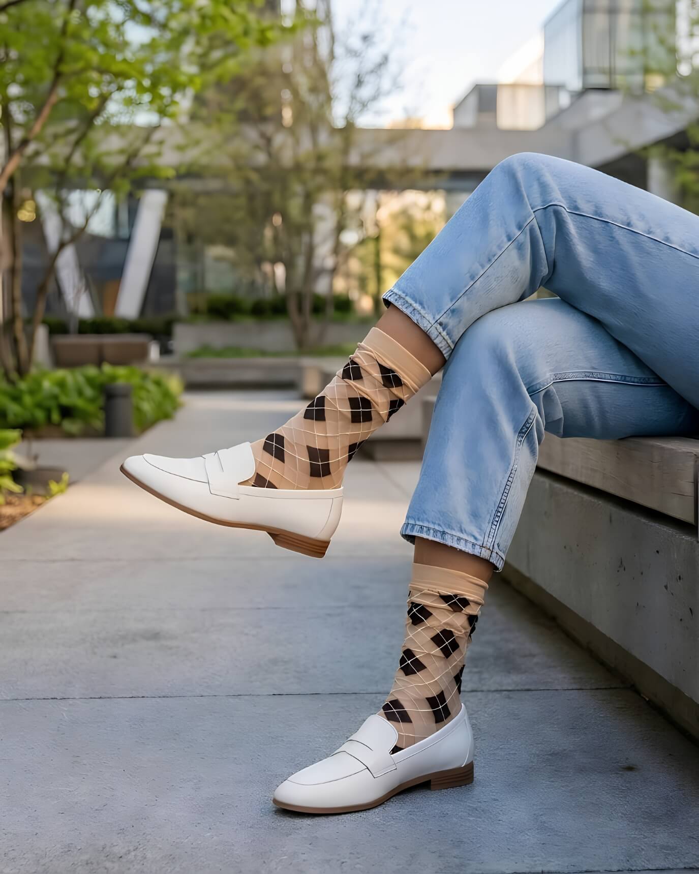 Sheer Argyle Crew Socks in beige and black featuring a soft diamond pattern, paired with white loafers and cuffed jeans while sitting on a concrete bench, showcasing the lightweight, sheer texture and casual styling.