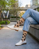 Sheer Argyle Crew Socks in beige and black featuring a soft diamond pattern, paired with white loafers and cuffed jeans while sitting on a concrete bench, showcasing the lightweight, sheer texture and casual styling.