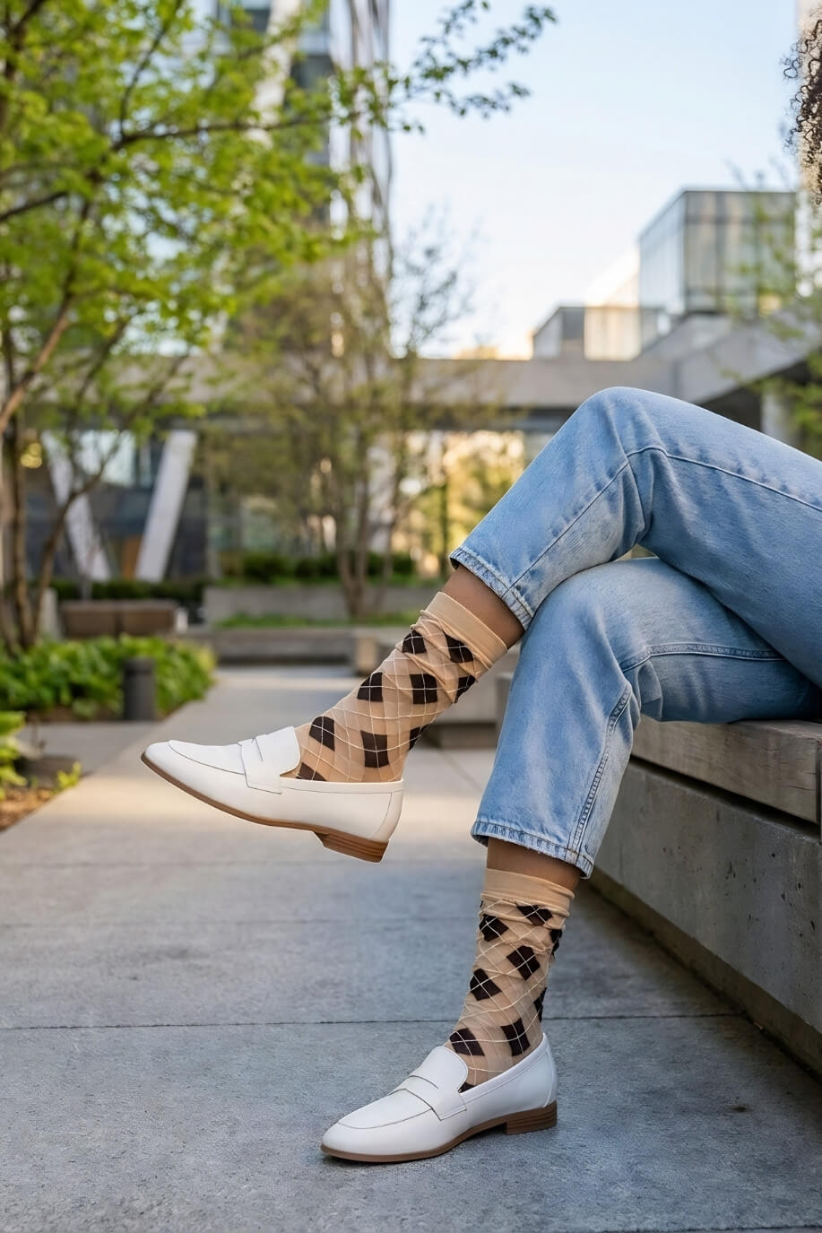 Sheer Argyle Crew Socks in beige and black featuring a soft diamond pattern, paired with white loafers and cuffed jeans while sitting on a concrete bench, showcasing the lightweight, sheer texture and casual styling.