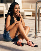 Shorty Socks, Wine Down featuring a pink ankle-length base with wine-themed artwork, dark toe and heel panels, and script text on the sole, worn while sitting on a sunny pool deck, highlighting relaxed lounge wear.