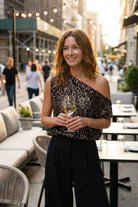 Premiere Night Top one shoulder sequin top styled on a city street restaurant setting with soft lights, paired with high waist trousers, highlighting the flattering drape and effortless evening wear feel.