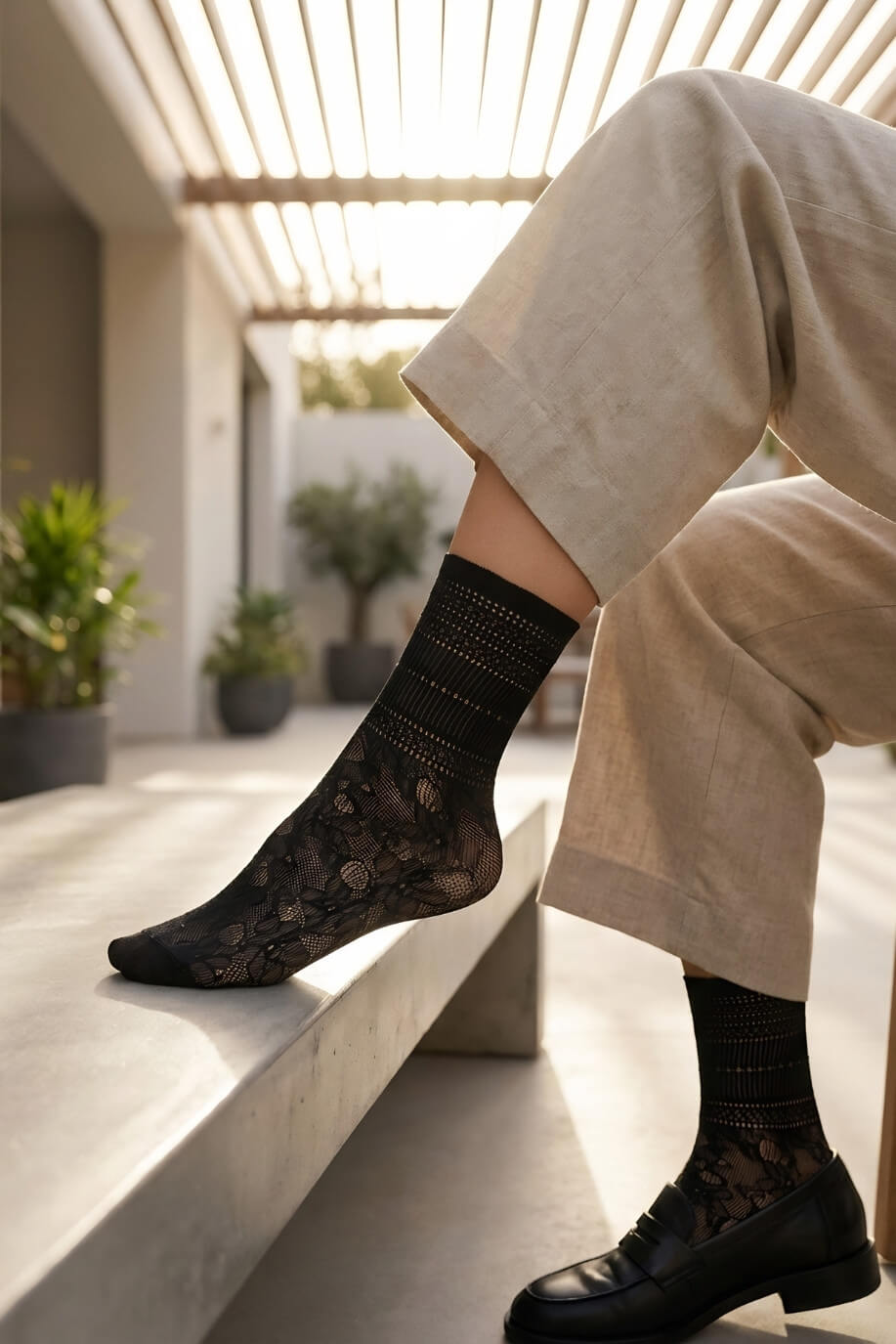 Sheer Floral Lace Crew Socks in black with intricate floral lace design, worn with tailored trousers and black loafers while sitting on a sunlit bench, showcasing the lightweight, sheer finish and elegant pattern.