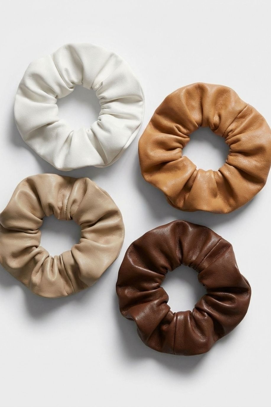 Set of four faux leather scrunchies in earthy neutral shades displayed in a grid layout, highlighting the smooth texture and structured gathered shape against a minimal white backdrop.