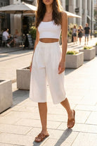 Bologna Bermuda Shorts, high-waist tailored knee-length shorts in off-white with front pleats, paired with a cropped white tank top in a city street setting, emphasizing the clean lines and relaxed, modern fit.