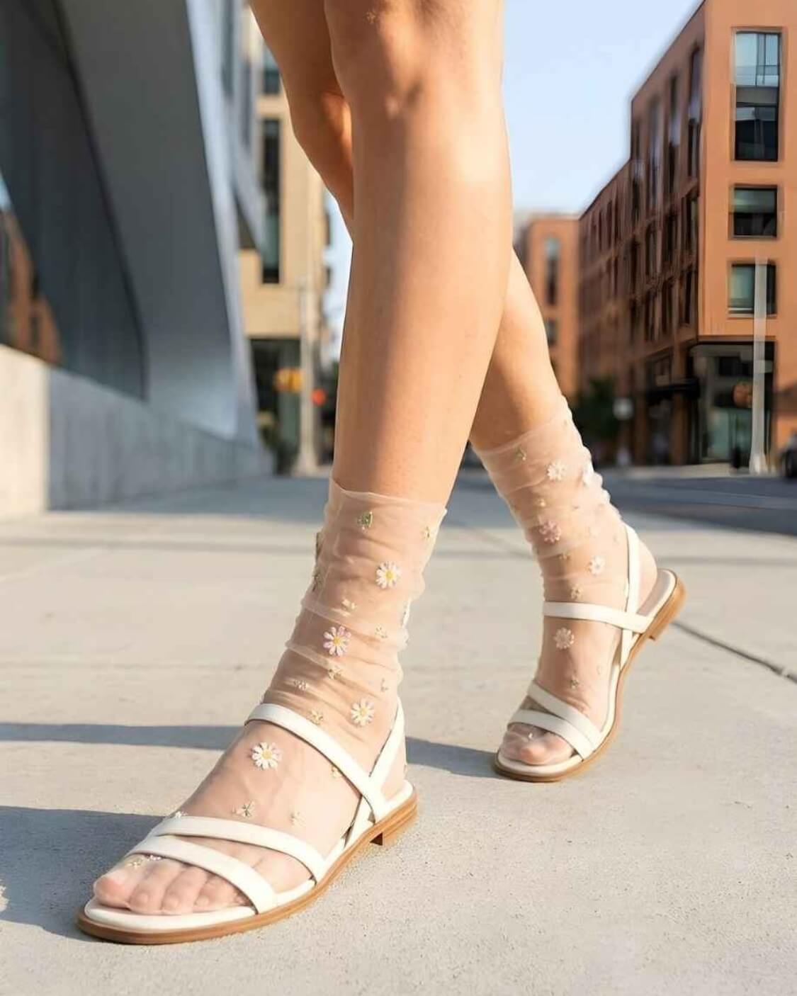 Sheer Garden Daisy Crew Socks in nude sheer fabric with floral embroidery, paired with white strappy sandals while walking on a city sidewalk.