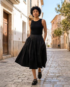 Poplin Cotton Smocked Flare Midi Skirt featuring a wide waistband, gathered flare silhouette, midi length, and side pockets, styled with a sleeveless top and loafers on a cobblestone street, highlighting everyday polished styling.
