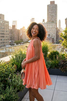 Accordion Pleated Dress, coral sleeveless pleated mini dress with a high neckline, shown from a side angle as the model stands on a rooftop garden, emphasizing the airy silhouette and soft drape of the pleats in motion.