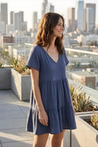 Cassady Dress featuring a soft, airy fit with a V-neckline, short sleeves, and tiered detailing, styled on a rooftop terrace with a blurred city skyline backdrop.