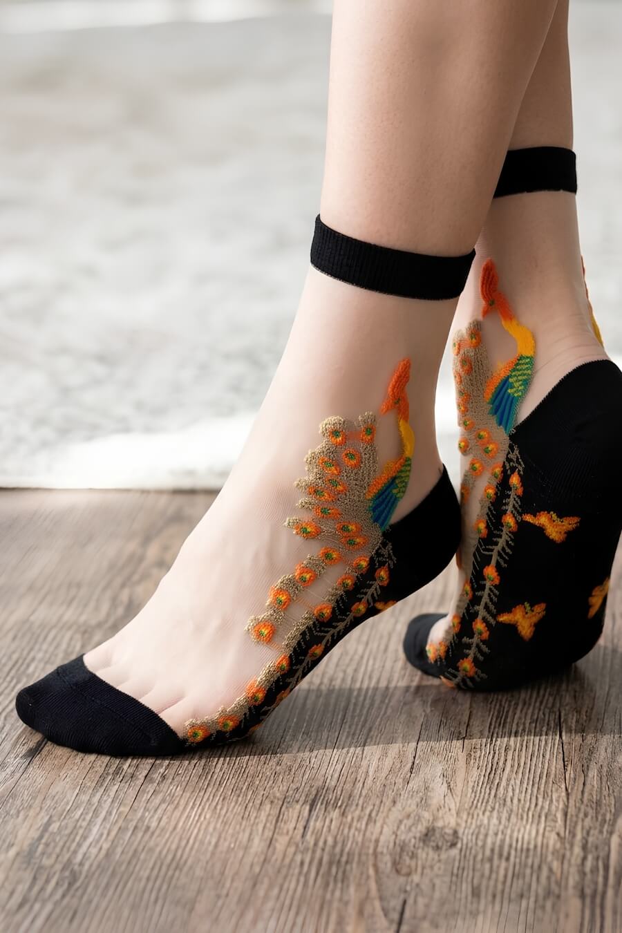 Peacock Sheer Mesh Socks featuring delicate nude-toned sheer mesh with colorful peacock embroidery and black reinforced toe, heel, and cuff trim, shown in a close-up standing pose on a wooden floor, emphasizing the lightweight texture, intricate stitching, and stylish feminine aesthetic.