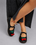 Cherry Accent Mary Jane Flats, black woven Mary Jane flats with red cherry embellishment and buckle closure, styled with a grey dress and statement anklet, highlighting close-up details and craftsmanship.