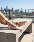 Roses Sheer Socks 2-Pack featuring sheer ankle socks with embroidered rose motifs and reinforced toe panels, worn while resting on a concrete bench in a bright city setting, highlighting the soft cuff fit and floral styling.