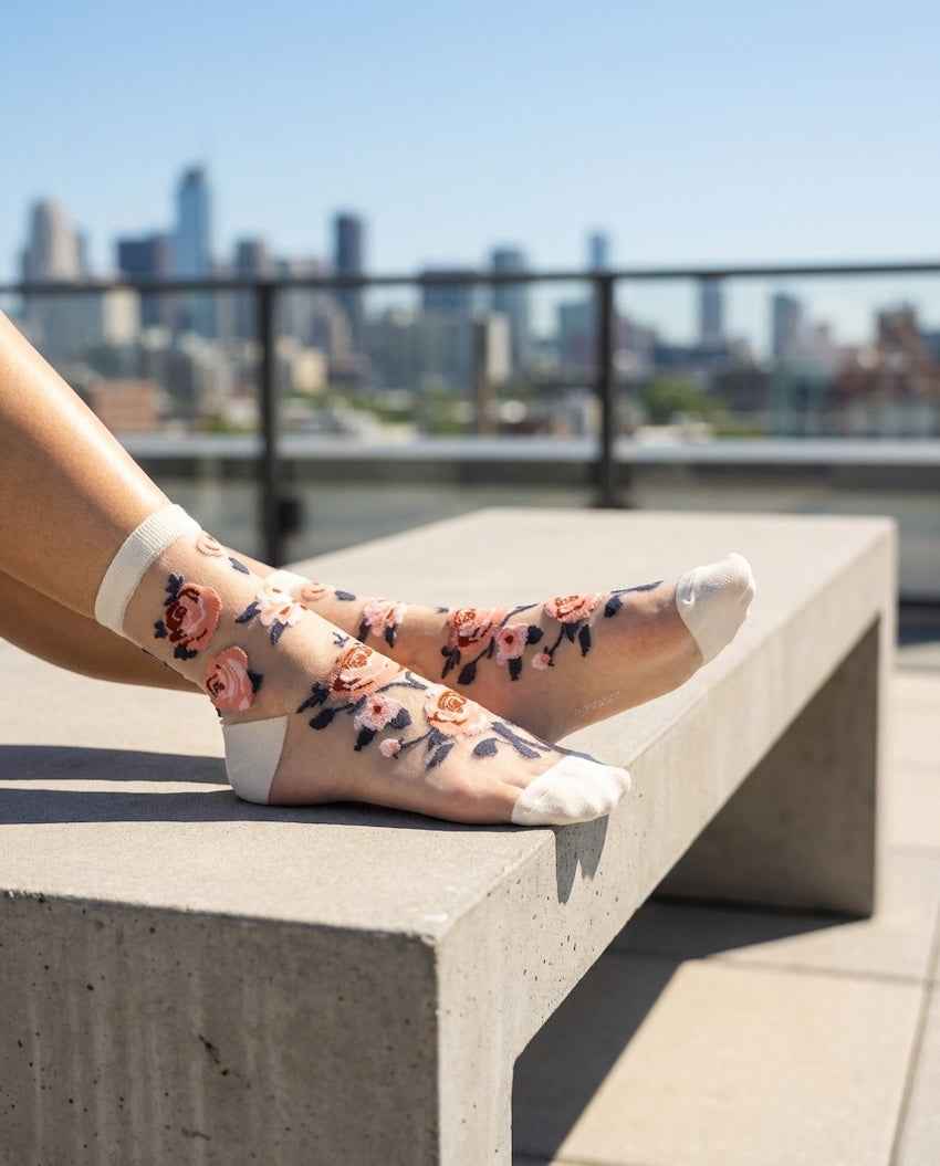 Roses Sheer Socks 2-Pack featuring sheer ankle socks with embroidered rose motifs and reinforced toe panels, worn while resting on a concrete bench in a bright city setting, highlighting the soft cuff fit and floral styling.