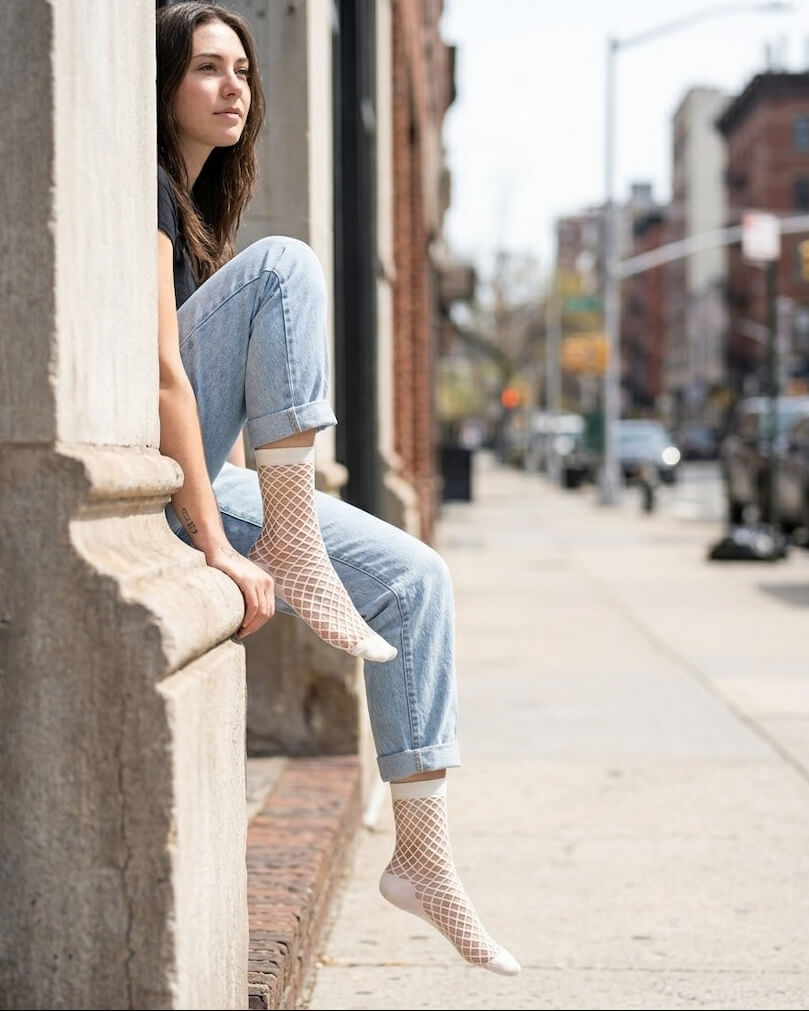 Midnight Lattice Fishnet Socks featuring an open mesh fishnet design with flexible stretch construction and solid cuff trim, worn with rolled denim jeans while seated on a city sidewalk ledge, highlighting breathable comfort and contemporary streetwear styling.