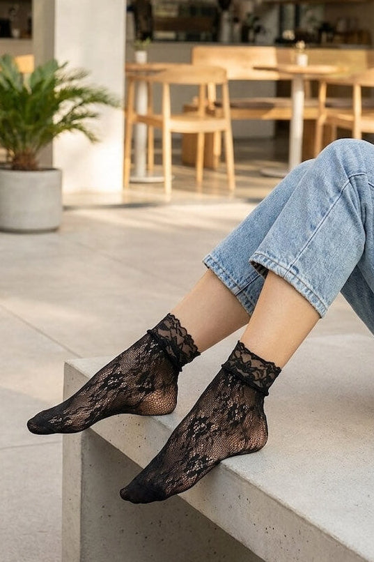 Romantic Floral Mesh Fishnet Socks in black with soft floral mesh pattern and scalloped cuff, worn alone with cuffed jeans while seated on a concrete ledge, emphasizing the breathable fishnet construction and romantic lace detail.