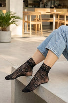 Romantic Floral Mesh Fishnet Socks in black with soft floral mesh pattern and scalloped cuff, worn alone with cuffed jeans while seated on a concrete ledge, emphasizing the breathable fishnet construction and romantic lace detail.