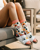 Crew Socks, Happy Camper featuring a full crew length with a pink cuff, camping graphics, and colorful outdoor motifs, worn with white buckle sandals while seated on patio furniture, highlighting the bold pattern and relaxed everyday wear.