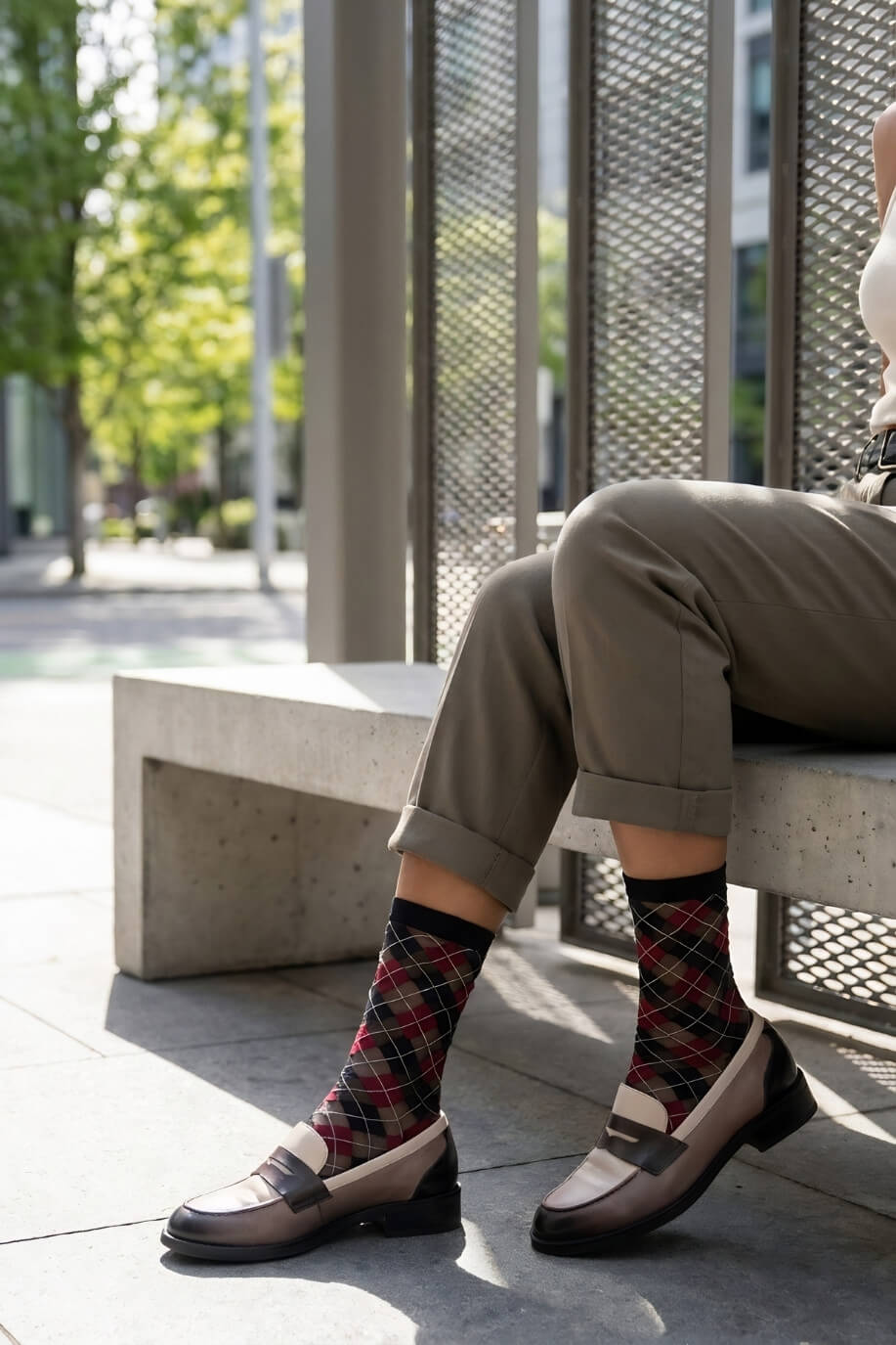 Sheer Argyle Crew Socks in black, red, and beige featuring a classic argyle motif, styled with rolled cuff pants while seated on a sunlit bench, highlighting the semi-sheer material and stylish patterned look.