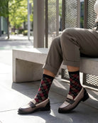 Sheer Argyle Crew Socks in black, red, and beige featuring a classic argyle motif, styled with rolled cuff pants while seated on a sunlit bench, highlighting the semi-sheer material and stylish patterned look.