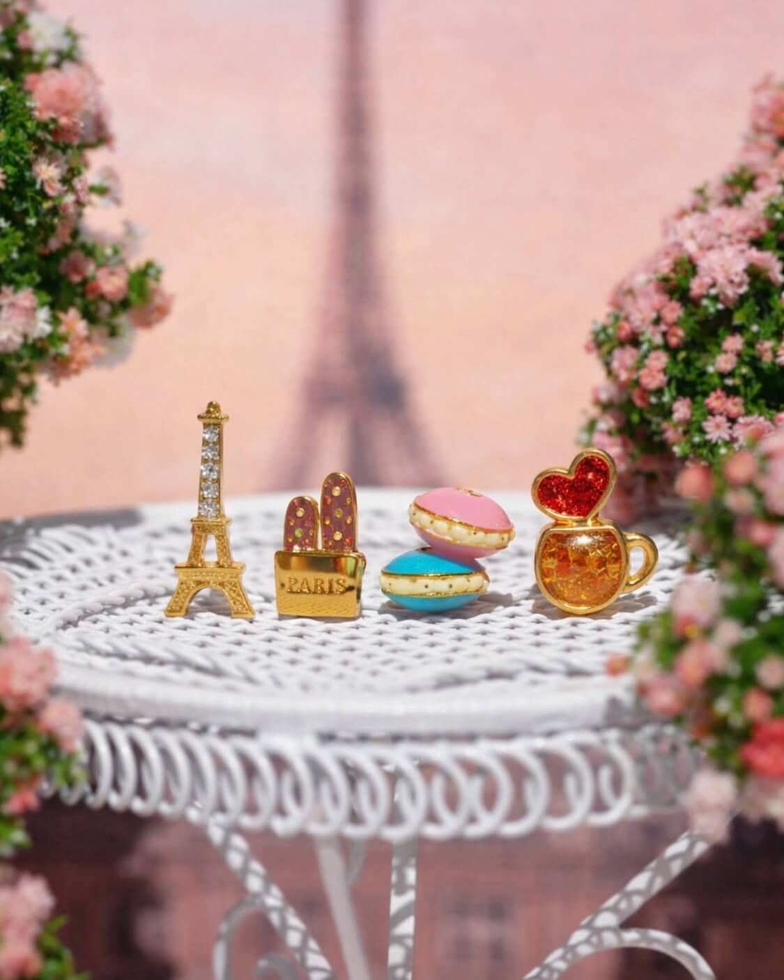 Café de Paris Stud Set, styled flat lay featuring Eiffel Tower, baguette bag, macarons, and coffee cup shaped studs arranged on a decorative table with a blurred Paris backdrop and floral accents.