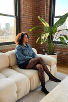 Fishnet Bow Tights featuring a delicate diamond fishnet pattern with bow details, styled with a denim jacket and black skirt while seated on a cream sofa.