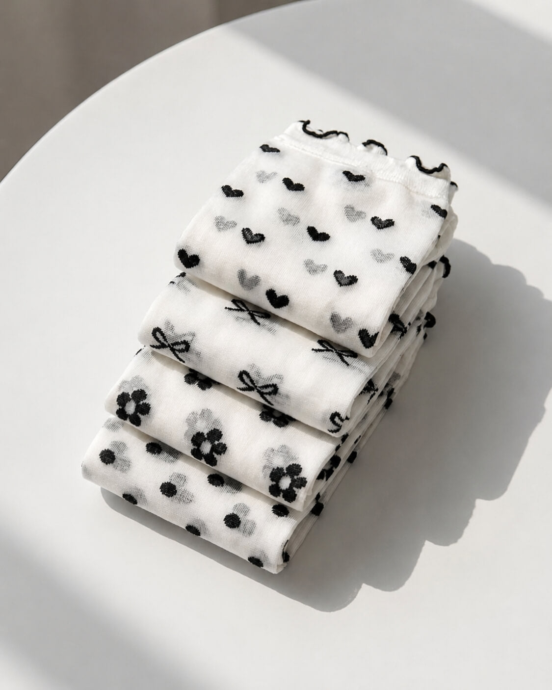 4-Pack Geo Print Ruffle Edge Sheer Socks featuring a coordinated collection of transparent mesh socks with geometric dot, heart, bow, and floral patterns, neatly folded and stacked on a minimalist surface to emphasize lightweight fabric, compact storage, and versatile everyday wear.